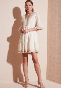 Robe blanche brodée avec un décolleté en V, des manches évasées et un ourlet à volants. Présente des finitions beiges et des motifs à jours sur l'ensemble.