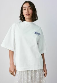 T-shirt en coton blanc à manches courtes, avec un logo "Pepsi-Cola" bleu sur la poitrine gauche et un col rond.