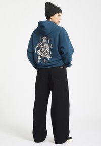 Niebieska bluza z kapturem z grafiką na plecach, połączona z czarnymi spodniami o szerokich nogawkach. Model nosi czapkę beanie w kolorze czarnym.