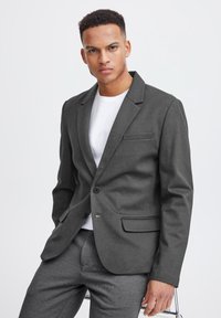 Grauer Blazer mit schmaler Passform, Ein-Knopf-Verschluss, Reverskragen und Brusttasche. Über einem weißen T-Shirt getragen, kombiniert mit grauen Hosen.