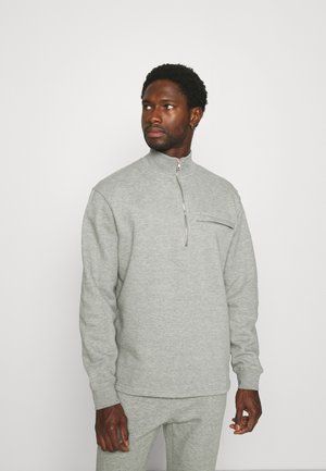 Lys grå pullover sweatshirt med høj krave, halvlukning og en brystlomme med en sort lynlås. Fremstillet af et blødt, tekstureret stof.