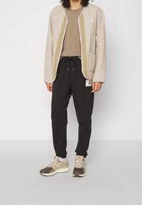Beige kviltad jacka med grön insida, grå t-shirt, svarta sweatpants och beige sneakers med grå detaljer och texturerade sulor.