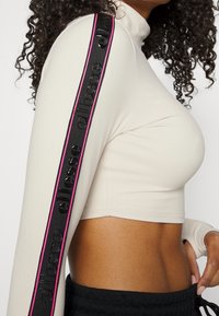 Widok z boku osoby noszącej beżowy krótki top z długim rękawem, z czarnym i różowym paskiem z logo na rękawie oraz czarne spodnie z sznurkiem.