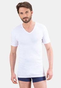 T-shirt blanc à manches courtes et col en V, fabriqué en tissu doux, avec une coupe ajustée et un col simple. Porté par-dessus un short de boxeur bleu foncé.
