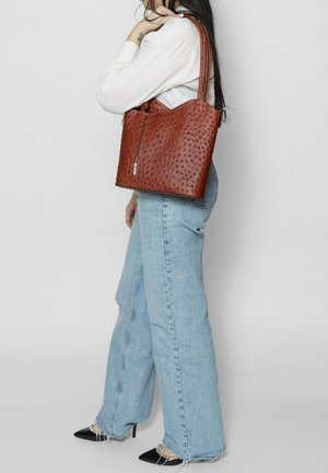 Sac tote en cuir marron avec motif texturé, fermeture éclair et double poignées. Porté avec un jean ample bleu clair et des talons noirs.