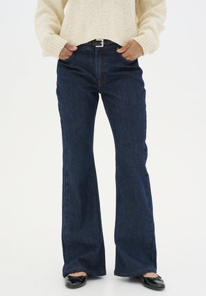 Dark blue flared jeans in denim material, featuring a mid-rise waist, five pockets, and contrasting stitching. Worn with a light sweater.