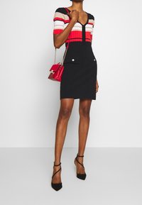 Robe noire courte avec un corsage ajusté, manches courtes rayées en rouge, beige et or. Présente un décolleté en V et deux poches décoratives à l'avant.