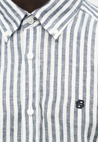Ριγέ πουκάμισο με κάθετες λευκές και ναυτικομπλε γραμμές, λευκά κουμπιά και μικρό ναυτικομπλε κεντημένο λογότυπο στο στήθος.