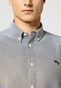 Camisa de botón en tela gris claro con un acabado texturizado. Presenta botones blancos y un logo bordado en el lado izquierdo del pecho.