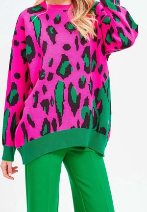 Woman wearing a vibrant pink sweater with green and black leopard print and green pants, posing with one hand near her head.