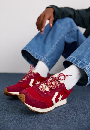 Rote Wildleder- und Mesh-Sneaker mit weißen Sternakzenten, reflektierenden Details und gemusterten rot-weißen Schnürsenkeln, kombiniert mit weißen Socken und blauen Jeans.