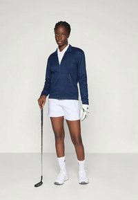 Mornarica zadrga jakna, bela polo majica in bele kratke hlače. Golf rokavica na levi roki, drži golf palico. Bele športne čevlje in nogavice.