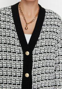 Schwarz-weiß gemusterte Strickjacke mit strukturiertem Strickmuster, ausgestattet mit einem schwarzen Kragen und drei goldfarbenen Knöpfen an der Vorderseite.