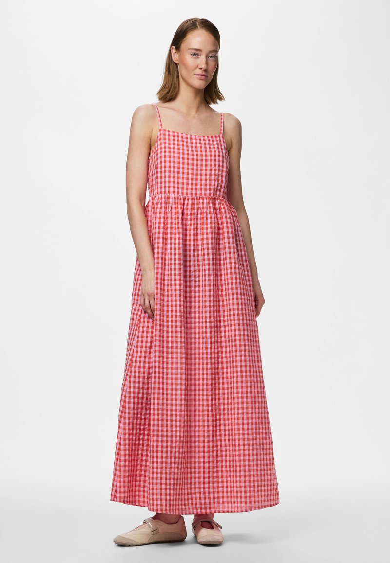 Femme portant une robe longue vichy rouge et blanche avec des fines bretelles et des chaussures plates beiges, debout contre un fond blanc uni.