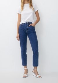 Blue high-waisted denim jeans with a straight leg fit, featuring five pockets and a front button closure, paired with a plain white t-shirt and wedges.