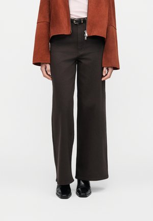 Person trägt schwarze Hose mit weitem Bein, schwarzen Gürtel, rostfarbene Wildlederjacke, weißes Oberteil und schwarze Stiefel mit quadratischer Spitze, steht vor weißem Hintergrund.