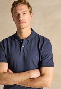 Tumesinine polo särk, millel on krae ja kolme nupuga sulgemine. Omab väikest helesinist logo. Sile kangas lühikeste varrukatega ja regulaarse lõikega.