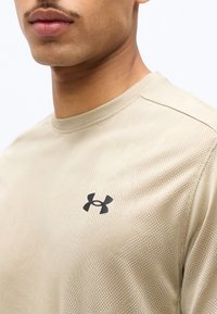 Κοντινό πλάνο ενός ατόμου που φοράει μπεζ υφασμένη μπλούζα Under Armour με μαύρο λογότυπο στο στήθος, σε λευκό φόντο.