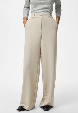 Beige viddebukser med høj talje og forlommer, lukning med knap og lynlås, kombineret med sorte spidse sko.