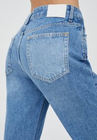 Jean en denim bleu clair à taille haute, avec deux poches arrière et un patch en cuir blanc à la ceinture. Texture lisse.