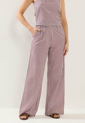 Tuta senza maniche con motivo a quadretti rosso e bianco, con pantaloni ampi, indossata con sandali marroni aperti sulle dita dei piedi, una mano in tasca.