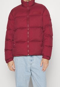 Bordeaux pufferjack met een hoge kraag, ritssluiting en gestikte secties. Voorzien van zijzakken en een logo op de borst.