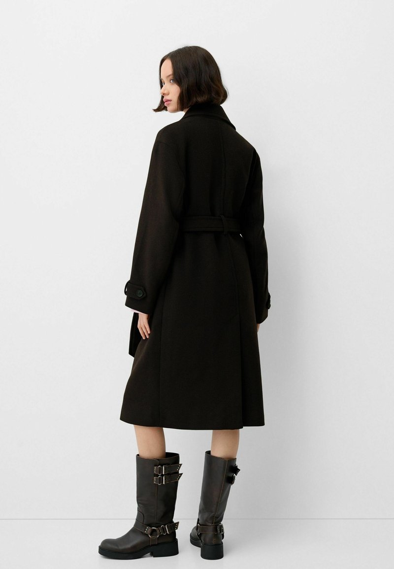 Czarny wełniany płaszcz z paskiem w talii, szerokim kołnierzem i guzikami na mankietach. Połączony z brązowymi skórzanymi botkami do kolan z klamrami.