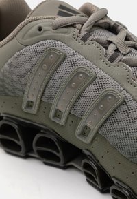 Athletic shoe with a grey mesh upper, green accents, and distinctive tubing sole. Features layered design with prominent lacing system.