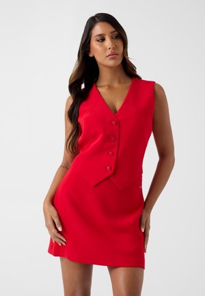 Kobieta ubrana w czerwoną bezrękawną sukienkę kamizelkę zapinaną na guziki, stojąca z rękami na biodrach, patrząca w bok na jednolitym tle.