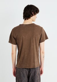 Brun bomull t-skjorte med en myk, falmet tekstur, korte ermer og rund hals, som viser svake tegn på slitasje på stoffoverflaten.