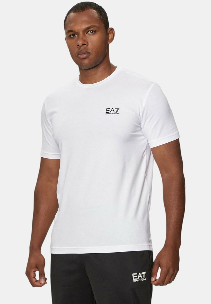 T-shirt bianco a maniche corte con collo rotondo, caratterizzato da un piccolo logo EA7 nero sul lato sinistro del petto. Materiale in cotone liscio, design aderente.