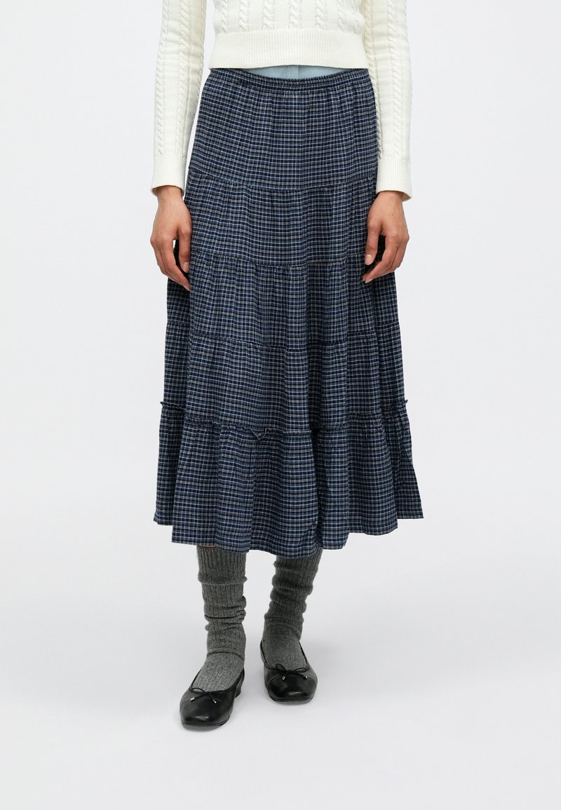 Spódnica w granatową kratę o warstwowym kroju, wykonana z lekkiego materiału. Charakteryzuje się rozkloszowanym kształtem, fakturowaną powierzchnią, zestawiona z szarymi, sięgającymi kolan skarpetami i czarnymi butami.