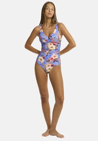 Das Model posiert selbstbewusst in einem blau gemusterten Badeanzug mit Blumenmuster, der die lebhaften Farben und die elegante Silhouette des Designs hervorhebt.