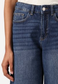 Jeans de mezclilla azul con un corte relajado, cinco bolsillos y cierre de botón. La tela presenta un leve desgaste, con detalles de costura en contraste.