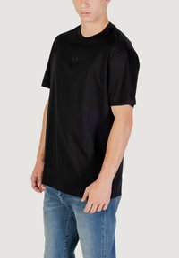 T-shirt nera a maniche corte in cotone, con un discreto patch del marchio sul petto, abbinata a jeans in denim azzurro chiaro.