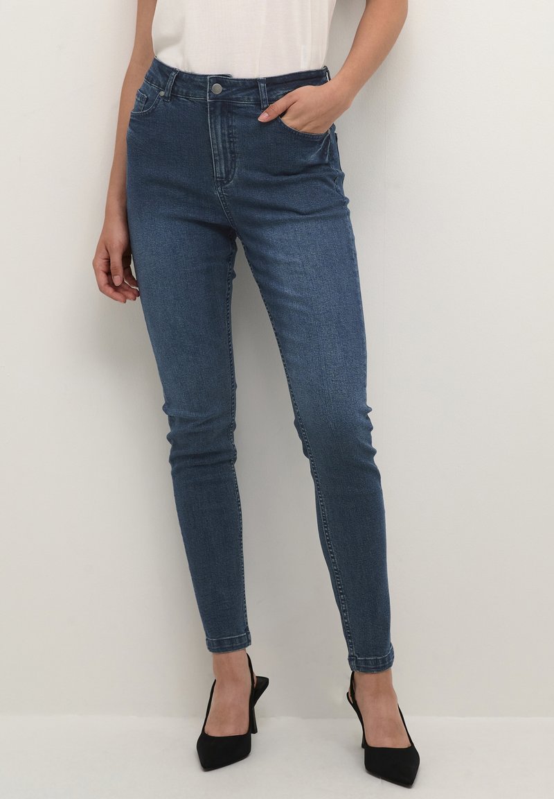 Femme portant un jean skinny bleu foncé ajusté, un haut blanc rentré et des talons pointus noirs, debout contre un mur clair uni.
