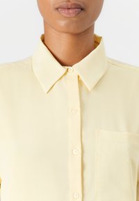 Camicia gialla con colletto classico, texture liscia e taschino sul petto. Presenta una lucentezza sottile e cinque bottoni abbinati.