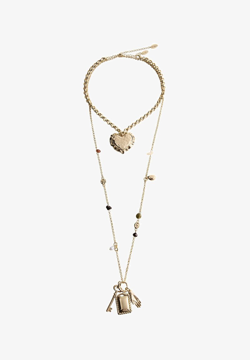 Collier doré superposé avec une chaîne épaisse et un pendentif en forme de cœur, et une chaîne plus longue avec de petites perles et des breloques en forme de clé, cadre et main.