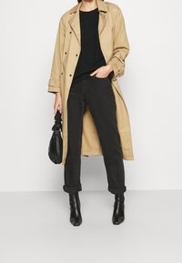 Trench-coat beige sur un pull noir en maille torsadée, associé à un jean noir retroussé et des bottines à talons noires ; tenant un petit sac noir.