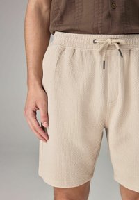 Beige teksturerede shorts med elastisk talje, snoretræk og sidelommer, prydet med et vævet mønster for ekstra dimension.