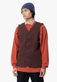 Bruine gewatteerde vest met een knopenvoer, gedragen over een oranje langemouwshirt, aangevuld met een blauwe gebreide beanie.