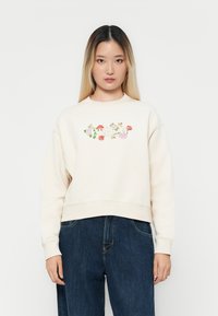 Felpa beige con ricami floreali in rosa e rosso. Presenta un collo a giro e polsini a costine, abbinata a pantaloni in denim scuro.