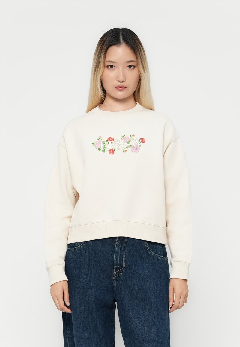 Felpa beige con ricami floreali in rosa e rosso. Presenta un collo a giro e polsini a costine, abbinata a pantaloni in denim scuro.