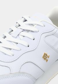 Hvite sneakers med hvite snørebånd, sydd detaljer og et lite gullsymbol på siden nær snøreområdet.