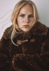 Brun, fluffig fuskpälsjacka med hög krage, försedd med spänne av läder och mjuk textur. Modellens hår flyter runt jackan.