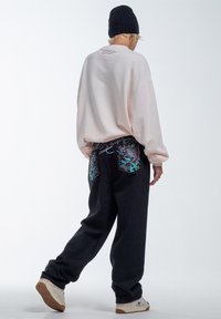 Sudadera de gran tamaño color rosa claro con detalles bordados en la parte posterior, combinada con pantalones de mezclilla oscuros que tienen diseños coloridos de peces en los bolsillos.