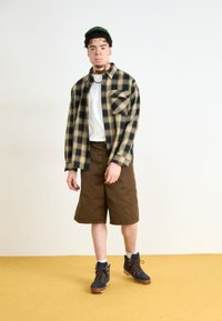 Cămașă de flanel cu mânecă lungă în carouri negre și bej, tricou alb, pantaloni scurți maro și cizme bleumarin cu accente galbene, amplasate pe un podea galbenă.