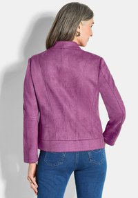 Taillierte lila Wildlederjacke mit hohem Kragen, langen Ärmeln und vertikalen Nahtdetails; kombiniert mit dunkelblauen Jeans.