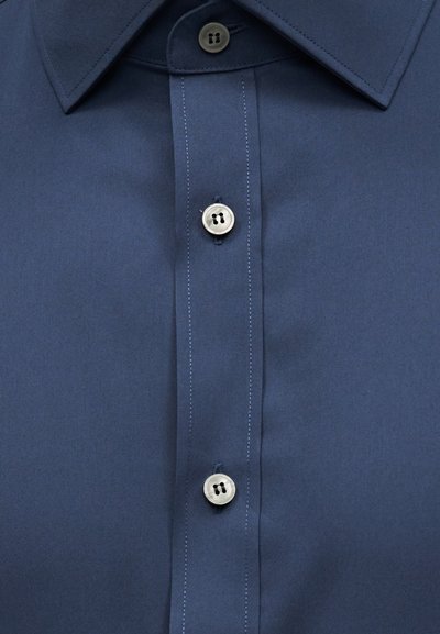 Σκούρο μπλε πουκάμισο μπροστά με μυτερό γιακά και τρία λευκά κουμπιά κατά μήκος της πατιλέτας, δύο κουμπωμένα και ένα ξεκουμπωμένο.
