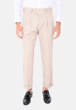 Pantaloni beige su misura con doppia piega frontale, chiusura a due bottoni, orli arrotolati e una superficie liscia, abbinati a mocassini neri.